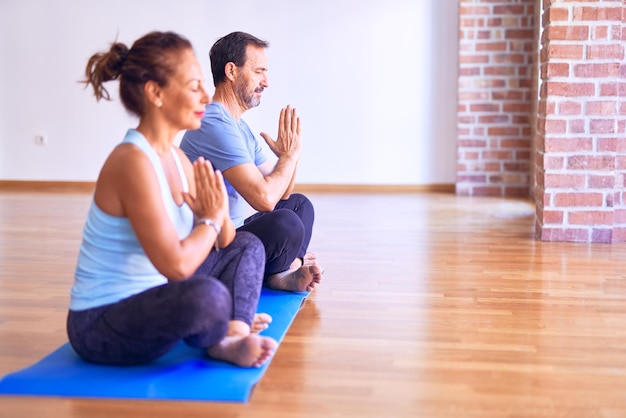 Beau couple sportif d'âge moyen assis sur un tapis pratiquant le yoga faisant la pose de prière au gymnase