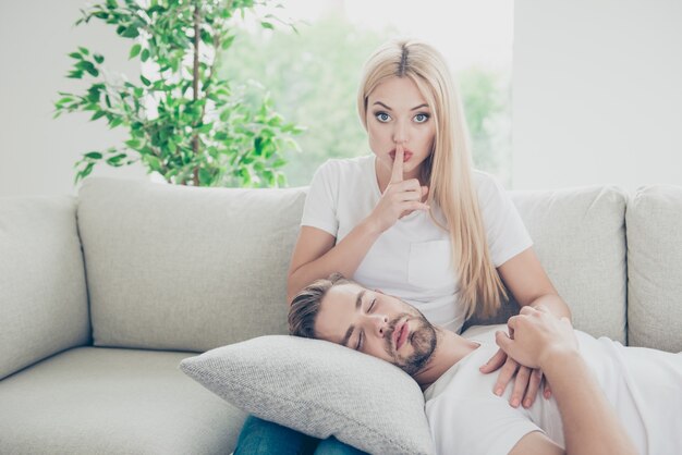 Beau couple en t-shirts blancs dans la maison fille s'asseoir sur l'homme divan dormant sur l'oreiller