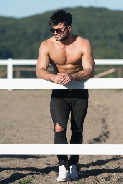 Beau cowboy homme debout fort et posant au ranch à l'extérieur portant un jean noir sur fond de ciel et de montagnes