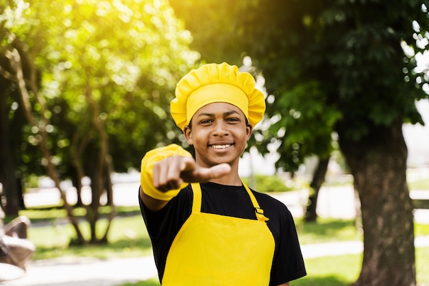 Beau cuisinier adolescent africain pointe vers vous Enfant noir cuisinier en toque de chef et uniforme de tablier jaune souriant et pointant vers vous à l'extérieur Publicité créative pour café ou restaurant