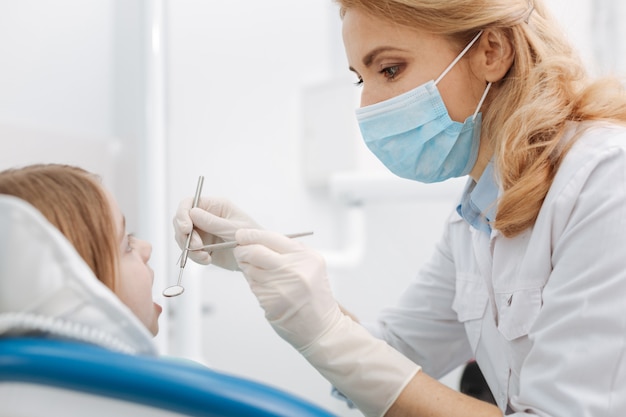 Beau dentiste pédiatrique expérimenté agissant très soigneusement tout en faisant son travail et en effectuant un examen des dents de ses patients