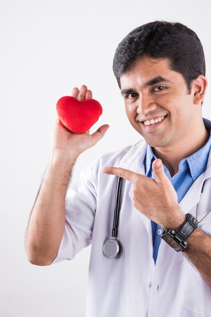 Beau docteur indien tenant un coeur en peluche rouge comme concept de soins cardiaques. debout isolé sur fond bleu