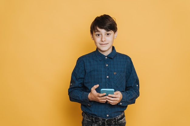 Beau écolier mignon souriant, portant une chemise décontractée et un jean tenant son téléphone portable, l'air confiant, sur fond jaune. Il a eu de bonnes nouvelles.