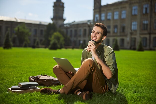 Beau étudiant rêveur avec une tasse de café en papier jetable regardant au loin