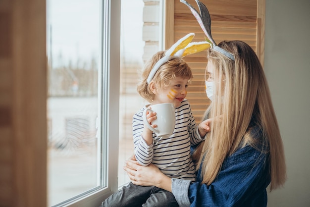 Photo beau fils et mère heureux dans des bandeaux avec des oreilles de lapin décorent des œufs avec un pinceau et des peintures lumineuses se préparent à célébrer pâques s'amuser à la maison