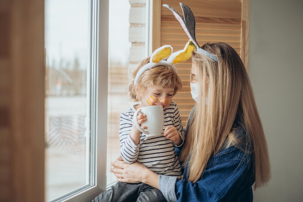 Photo beau fils et mère heureux dans des bandeaux avec des oreilles de lapin décorent des œufs avec un pinceau et des peintures lumineuses se préparent à célébrer pâques s'amuser à la maison