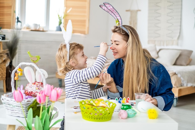 Beau fils et mère heureux dans des bandeaux avec des oreilles de lapin décorent des œufs avec un pinceau et des peintures lumineuses se préparent à célébrer Pâques s'amuser à la maison