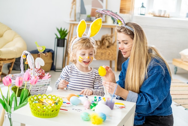 Beau fils et mère heureux dans des bandeaux avec des oreilles de lapin décorent des œufs avec un pinceau et des peintures lumineuses se préparent à célébrer Pâques s'amuser à la maison