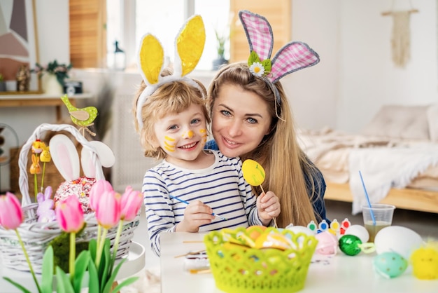 Photo beau fils et mère heureux dans des bandeaux avec des oreilles de lapin décorent des œufs avec un pinceau et des peintures lumineuses se préparent à célébrer pâques s'amuser à la maison