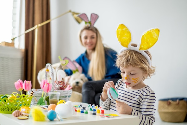 Photo beau fils et mère heureux dans des bandeaux avec des oreilles de lapin décorent des œufs avec un pinceau et des peintures lumineuses se préparent à célébrer pâques s'amuser à la maison