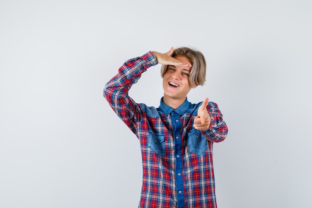 Beau garçon adolescent en chemise à carreaux pointant vers l'avant, avec la main sur la tête et l'air énergique, vue de face.