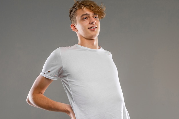 Beau garçon adolescent émotionnel qui pose en studio contre gris, mec dans un t-shirt gris avec un dos douloureux