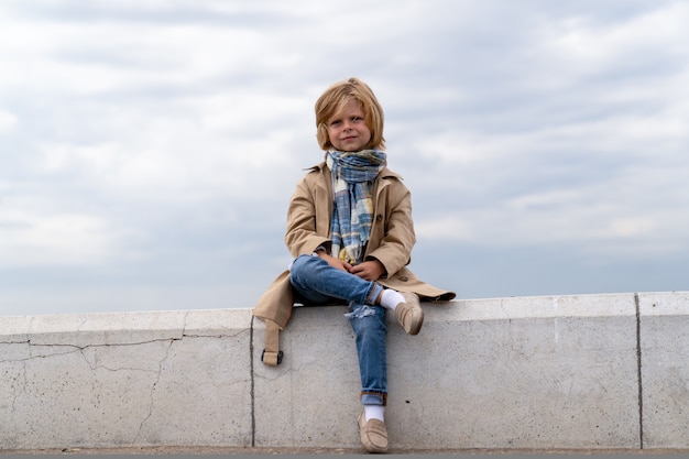 Un beau garçon blond de cinq ans dans un manteau et un foulard est assis sur un banc de pierre sur le remblai