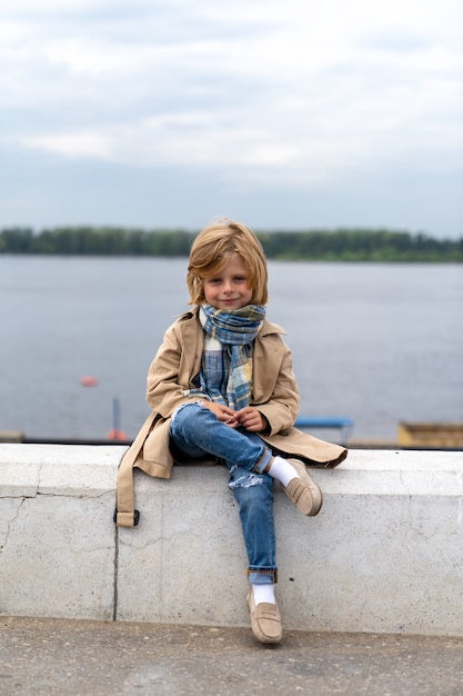 Photo un beau garçon blond de cinq ans dans un manteau et un foulard est assis sur un banc de pierre sur le remblai
