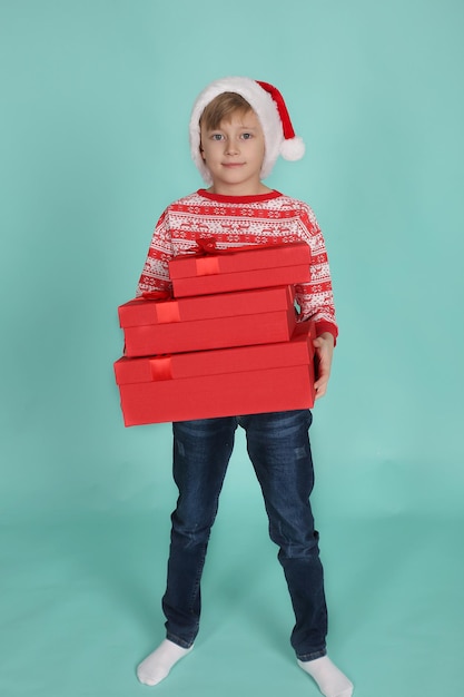 un beau garçon blond dans un pull de Noël chapeau de père Noël tient des coffrets cadeaux dans ses mains