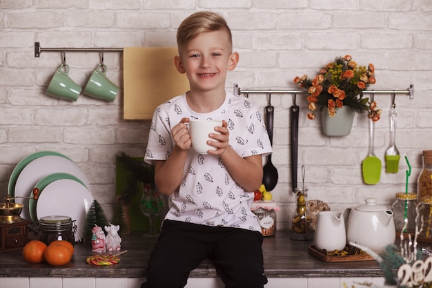 Un beau garçon blond est assis sur la table de la cuisine avec une grande tasse blanche à la main. Photo drôle