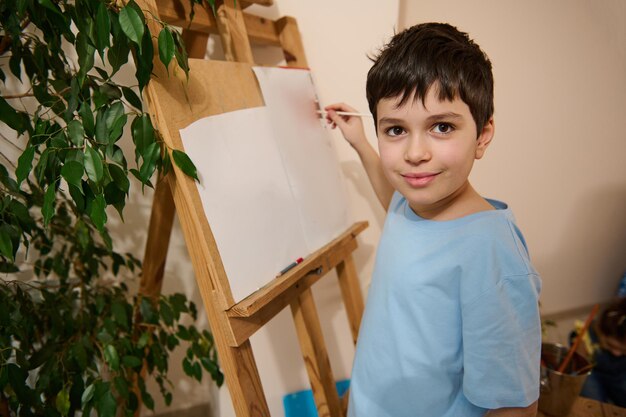 Beau garçon caucasien d'âge scolaire dessinant sur toile debout sur un chevalet en bois et profitant d'un cours d'art Développement de divertissement pour enfants et concept d'éducation créative artistique