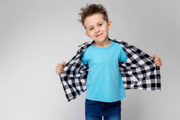 Un beau garçon dans une chemise à carreaux, une chemise bleue et un jean se dresse sur un fond gris