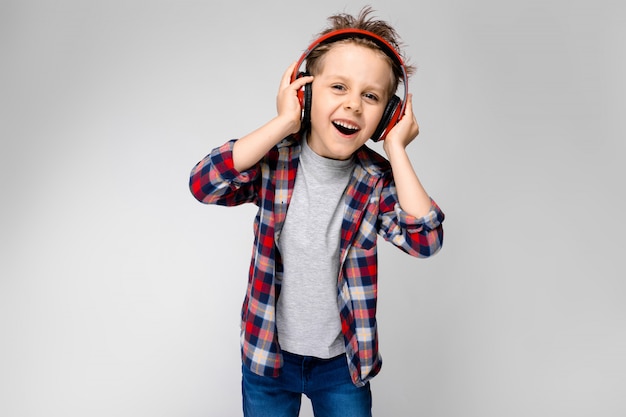 Un beau garçon dans une chemise à carreaux, une chemise grise et un jean se dresse sur un fond gris. Un garçon au casque rouge chante une chanson.