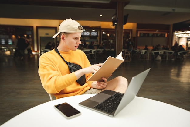 beau garçon étudiant en vêtements décontractés assis avec un ordinateur portable sur le bureau et un livre dans les mains au café