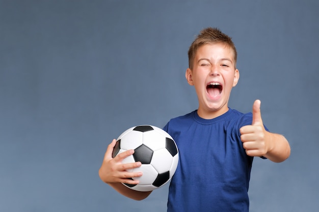 Beau garçon de fan caucasien hurlant tient le ballon de football et faisant des gestes les pouces vers le haut