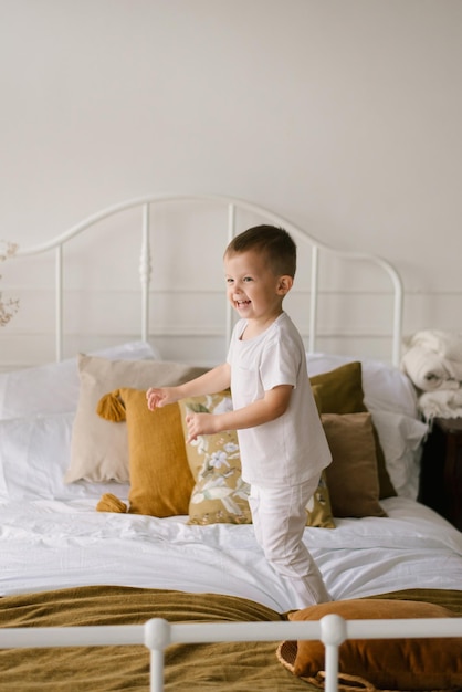 Beau garçon mignon de quatre ans en vêtements blancs sourit et saute sur le lit sur le fond clair de la maison