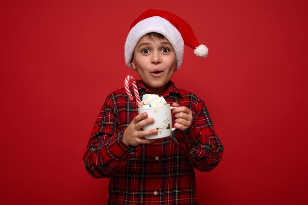 Beau garçon préadolescent étonné portant un bonnet de Noel pose sur fond rouge avec une tasse de boisson au chocolat chaud avec des guimauves et de la canne en bonbon à rayures. Concept de Noël avec espace de copie pour l'annonce