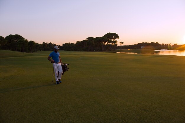 beau golfeur du moyen-orient portant un sac et marchant jusqu'au prochain trou du terrain de golf sur un magnifique coucher de soleil en arrière-plan