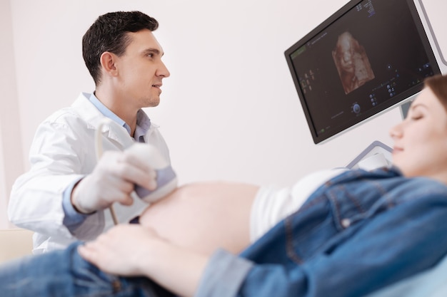 Beau gynécologue qualifié attentif travaillant tout en utilisant un équipement à ultrasons pour l'examen ultrasonique du ventre de femme enceinte et tenant une sonde linéaire