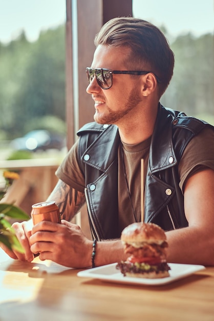 Un beau hipster avec une coupe de cheveux et une barbe élégantes est assis à une table, s'est arrêté pour déjeuner dans un café en bordure de route.