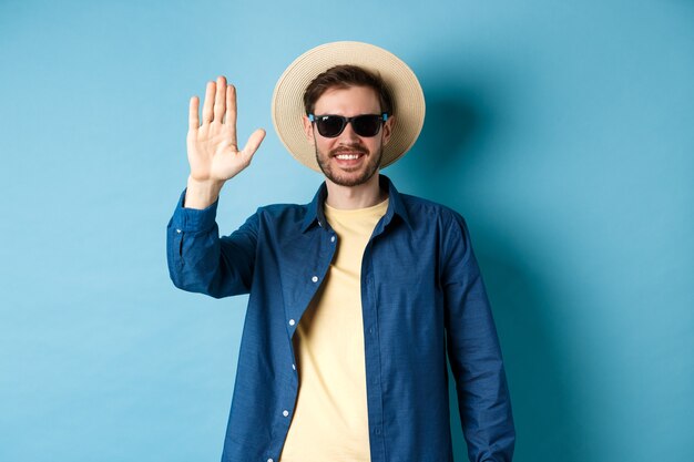 Beau hipster en vacances d'été, renonçant à la main et souriant, disant bonjour, portant des lunettes de soleil et un chapeau de paille, fond bleu.