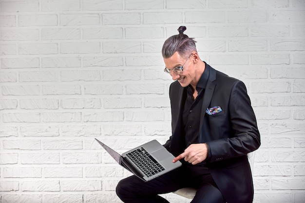 Beau homme d'affaires dans des lunettes et costume tenant ordinateur portable dans les mains et écrire quelque chose. Vue de côté. Sur brique blanche