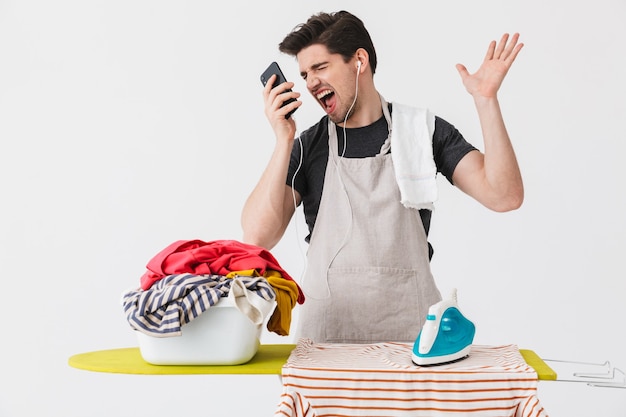 Beau homme de maison brune en colère portant un tablier debout isolé sur blanc, repassage des vêtements sur une planche, parlant au téléphone mobile