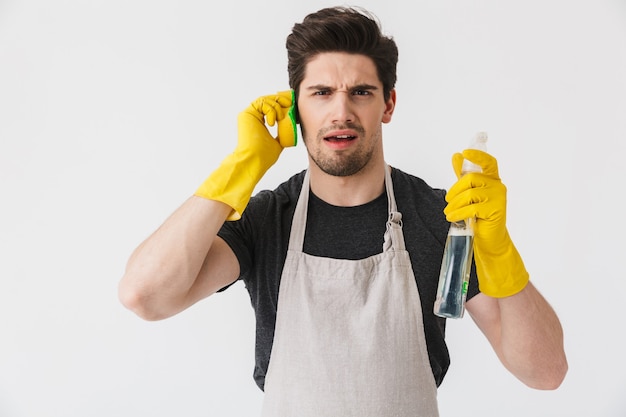 Photo beau homme de maison brune portant un tablier isolé sur blanc, faire semblant de parler au téléphone portable avec une éponge