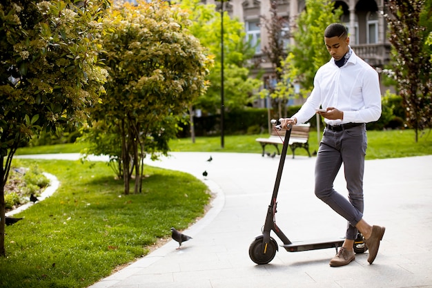 Beau jeune afro-américain à l'aide de téléphone mobile en se tenant debout par scooter électrique dans une rue