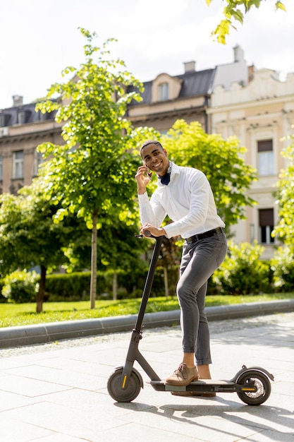 Beau jeune afro-américain utilisant un téléphone portable en se tenant debout avec un scooter électrique dans une rue