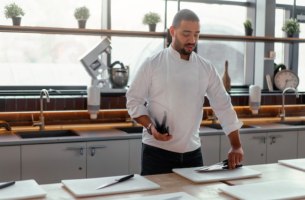 Beau jeune chef d'origine ethnique africaine pose des couteaux sur la table devant une master class culinaire. Homme afro en uniforme de chef se prépare pour les cours de cuisine.