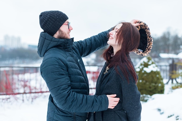 Beau jeune couple d'amants à la mode dans des vêtements d'hiver à la mode sur la rue beau gars met un chapeau sur sa petite amie et sourit s'amuser