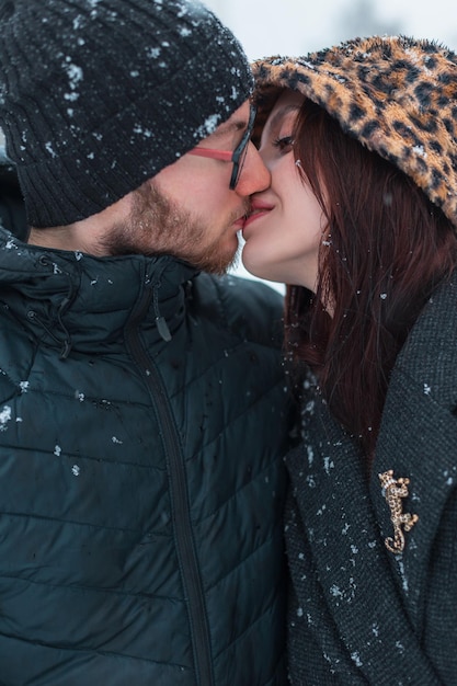 beau jeune couple d'amants à la mode vêtements d'hiver s'embrassent dans un parc avec la neige beau parka dans un chapeau tricoté et veste embrasse sa petite amie