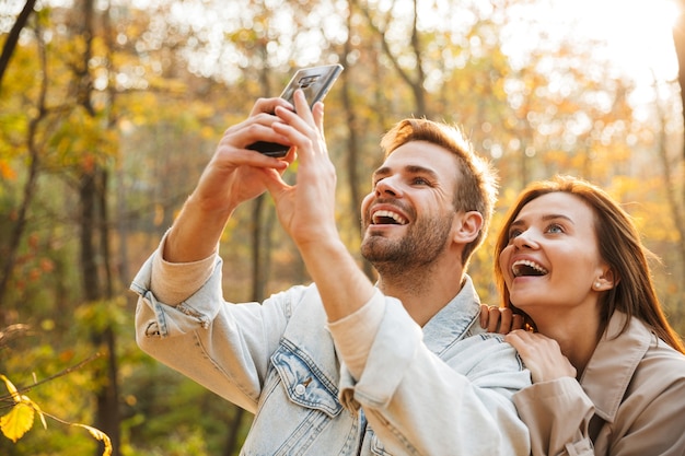 Beau jeune couple amoureux utilisant un téléphone portable tout en passant du temps au parc d'automne, en prenant un selfie