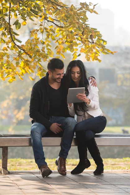 Beau jeune couple assis sur un banc dans le parc par une belle journée d'automne, ils utilisent Internet via une tablette numérique