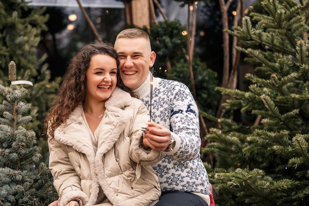 Un beau jeune couple assis près d'arbres de Noël en vêtements blancs marchant près de guirlandes Heureux homme et femme romance célébration de Noël amour amusant chandail blanc nouvel an rue à l'extérieur