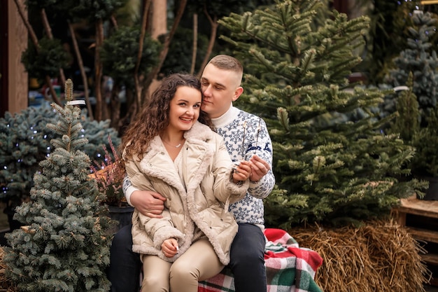 Un beau jeune couple assis près d'arbres de Noël en vêtements blancs marchant près de guirlandes Heureux homme et femme romance célébration de Noël amour amusant chandail blanc nouvel an rue à l'extérieur