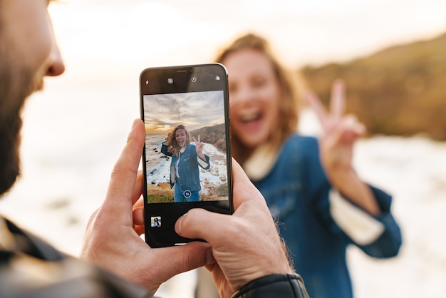 beau jeune couple caucasien prenant une photo sur un téléphone portable en marchant au bord de la mer