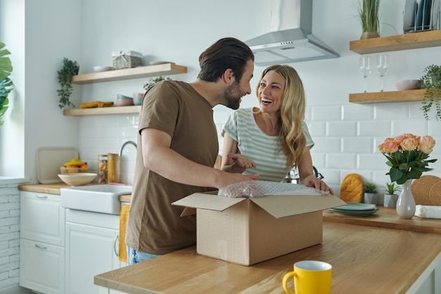 Un beau jeune couple déballe une boîte et sourit en se tenant debout dans la cuisine domestique.