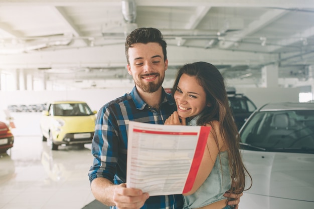 Beau jeune couple debout chez le concessionnaire en choisissant la voiture à acheter