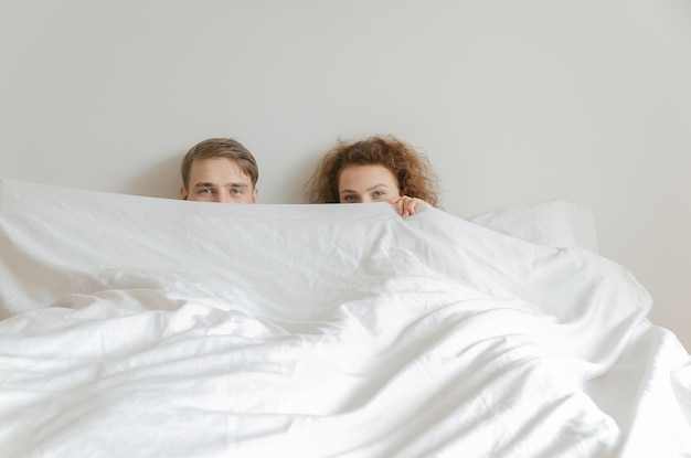 Un beau jeune couple a dormi ensemble, s'est couvert le visage avec une couverture et a regardé la caméra