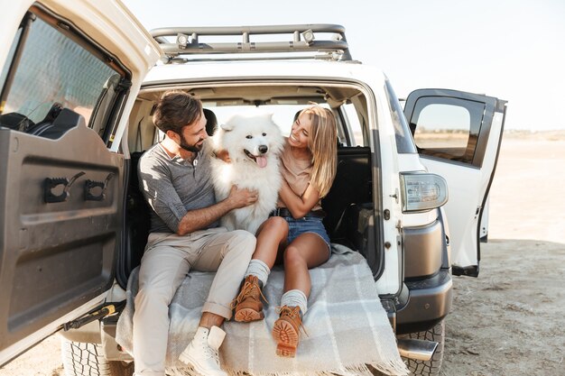 Beau jeune couple heureux assis à l'arrière de leur voiture à la plage, jouant avec un chien