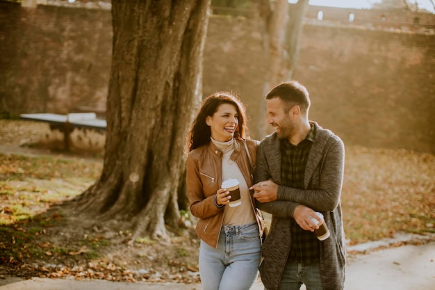 Beau jeune couple marchant dans un parc en automne avec du café pour aller des tasses dans les mains