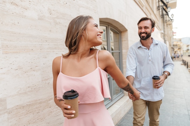 Beau Jeune Couple Marchant à L'extérieur Le Long De La Rue De La Ville, Se Tenant La Main, Buvant Du Café à Emporter